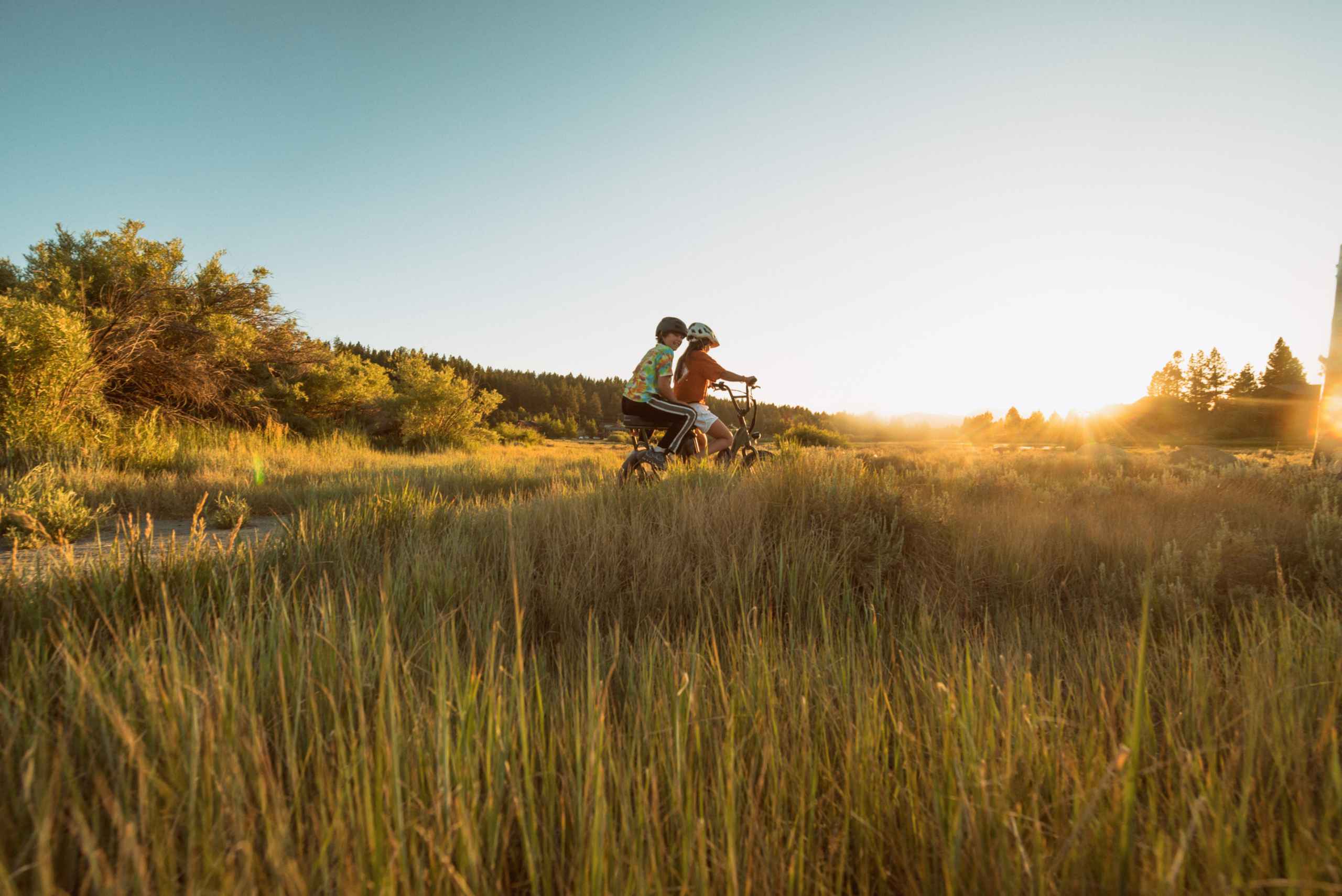 Two children riding a e-bike through a grassy field at sunset.